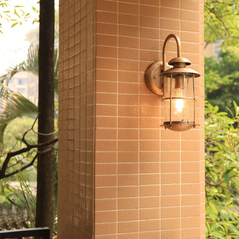 Rustic bronze cylindrical outdoor sconce with glass lantern attached to tiled column, surrounded by greenery.